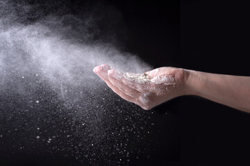 Fototapeta na wymiar Wheat flour flying in the wind with female hands on a black background, spraying white powder.