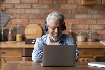 Grandpa speaking. Happy positive aged male in wireless headset sit by computer at kitchen table talk by video call with grandchild. Laughing old man enjoy chat with friend via conference app on laptop