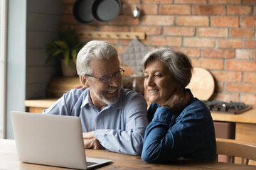 Fototapeta na wymiar Old age and modern tech. Retired family couple sit at kitchen table use laptop watch photo video read latest news online. Aged husband share discuss interesting information on website with senior wife