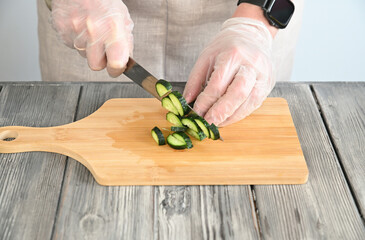 A woman in transparent gloves is cutting fresh cucumbers into round slices.