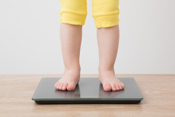 Baby girl with barefoot standing on weight scales on wooden floor at light gray wall background. Closeup. Front view. Care about body. Weight control concept.