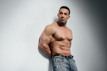 A young athlete bodybuilder poses in the studio topless in jeans near the wall. Sport.