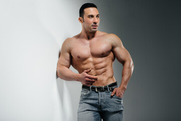 A young athlete bodybuilder poses in the studio topless in jeans near the wall. Sport.