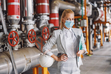 Middle-aged successful businesswoman in formal wear with protective face mask holding clipboard with documentation while standing in power plant during corona virus pandemic.