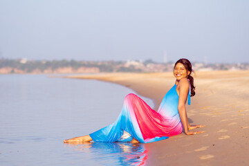 pretty young black woman having fun at the beach and smiling, wearing a colorful dress, sitting near the water