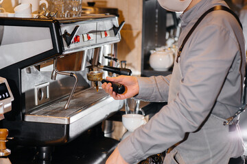 Close-up of a masked barista preparing a delicious coffee at the bar in a cafe. The work of...