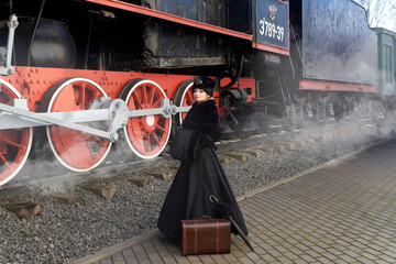 A beautiful girl in a historical retro dress against the background of an old steam locomotive at the station.
