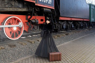 Fototapeta na wymiar A beautiful girl in a historical retro dress against the background of an old steam locomotive at the station.