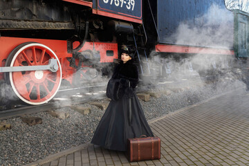 A beautiful girl in a historical retro dress against the background of an old steam locomotive at the station.