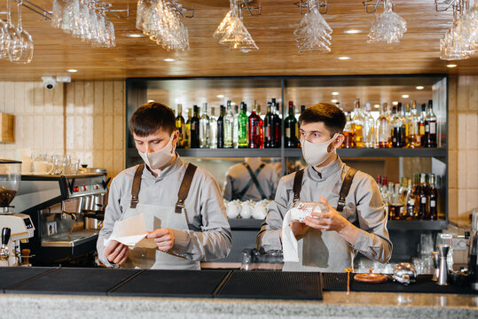 Two stylish bartenders in masks and uniforms during the pandemic, rub glasses to shine. The work of restaurants and cafes during the pandemic.