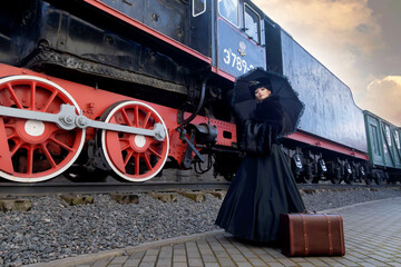 A beautiful girl in a historical retro dress against the background of an old steam locomotive at the station.