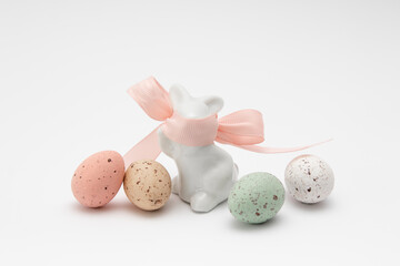 Four chocolate eggs, white, red, green and yellow with specks on a white background. Imitation of quail eggs. Porcelain rabbit with pink ribbon covering the mo
