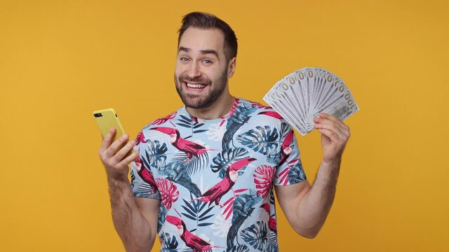 Surprised happy bearded young man 20s in casual basic summer t-shirt isolated on yellow background studio. People lifestyle concept. Using mobile cell phone hold fan of cash money in dollar banknotes
