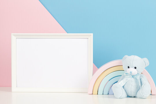 White Blank Frame With Teddy Bear And Pastel Toy Rainbow On White Desk. Baby Room Art Frame Mock Up With Baby Kid Toys Over Pink And Blue Background