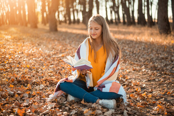 Young cute schoolgirl sitting in plaid in autumn forest and reading a book. Cheerful girl in park with a book