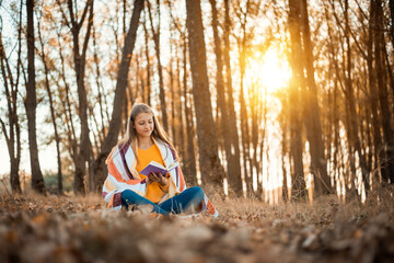 Portrait of a girl in an Autumn forest with a book against setting sun. Young girl reading a book in bright colorful forest and breathing fresh air