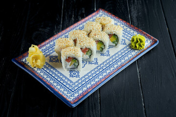 Appetizing Japanese sushi roll in sesame seeds with salmon, avocado and cream cheese in a blue plate on a black wooden background. Japanese cuisine. Philadelphia roll with sesame seeds