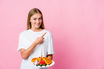 Young russian woman eating a waffle isolated smiling and pointing aside, showing something at blank space.