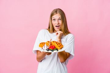 Young russian woman eating a waffle isolated praying for luck, amazed and opening mouth looking to front.