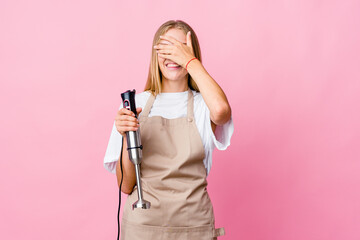 Young russian cook woman holding an electric mixer isolated covers eyes with hands, smiles broadly waiting for a surprise.