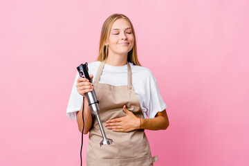Young russian cook woman holding an electric mixer isolated touches tummy, smiles gently, eating and satisfaction concept.