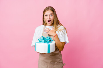 Young russian baker woman holding a delicious cake praying for luck, amazed and opening mouth looking to front.