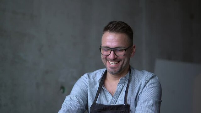 Bearded middle aged male artist in glasses sitting on a background of workplace. Linocut master in apron smiling and looking straight in camera and laughing. 4K footage
