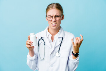 Young russian doctor woman holding pills bottle on blue upset screaming with tense hands.