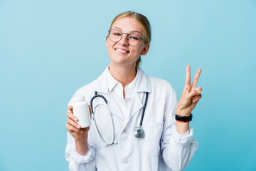 Young russian doctor woman holding pills bottle on blue showing number two with fingers.