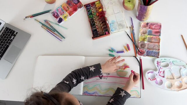 Cute little girl painting using watercolor paints. Top view of charming little girl painting using watercolors while sitting at the table in her room at home with notebook nearby. 4k footage