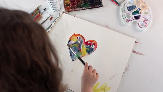 Small child is drawing colorful heart on white paper. Art school concept. Top view of a girl kid sitting at the table and painting with watercolor paints. 4k footage
