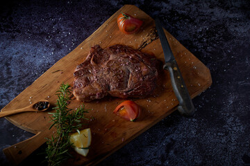 Grilled beef steak on wooden board with salt and other seasonings on a dark surface.
