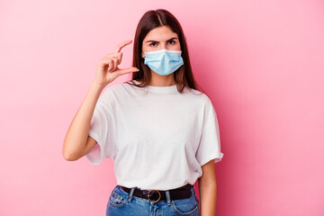 Young caucasian woman wearing a mask for virus isolated on pink background holding something little with forefingers, smiling and confident.