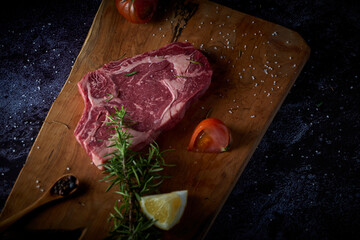 Fresh and raw beef steak on wooden board with salt and other seasonings on dark surface