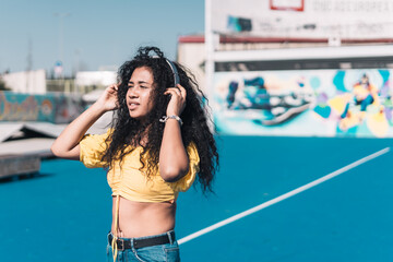 Young African American girl in a yellow t-shirt listening to music in an urban location looking at the sun.