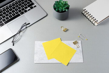 Grey workplace with blank yellow note paper, laptop, mobile phone, open notebook, glasses and succulent