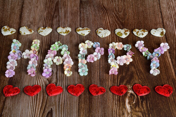 On a wooden background, the word HAPPY is spelled out in large letters of multicolored flowers. Decorative hearts are lined top and bottom in a line