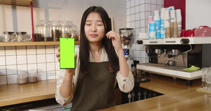 Cheerful Asian coffee shop owner holding smartphone with chroma key. Young female bartender standing behind bar and showing gree screen to camera in cafe. Business, carrer, job concept.
