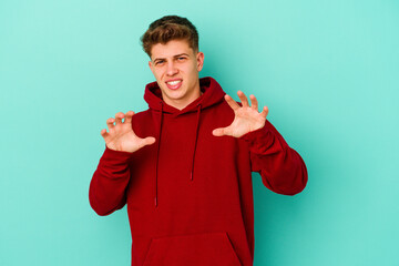 Young caucasian man isolated on blue background showing claws imitating a cat, aggressive gesture.