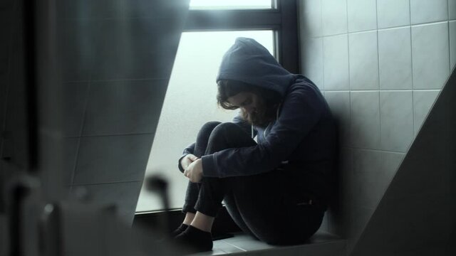 A depressed, sad and lonely young woman with hoody sitting in front of window. Moody and dark atmosphere of Millennial girl at home. Concept of depression, isolation, loneliness, mental health. 4K.