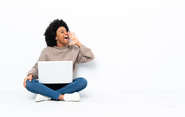 Young African American woman with a laptop sitting on the floor shouting with mouth wide open to the lateral