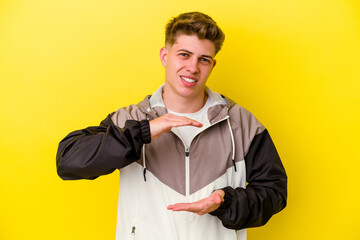 Young caucasian man isolated on yellow background holding something with both hands, product presentation.