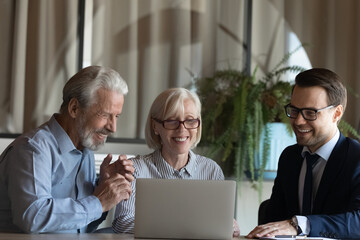 Smiling male realtor or broker consult middle-aged Caucasian couple buyers show project on laptop....