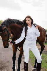 Foto op Canvas A young pretty girl rider poses near a thoroughbred stallion on a ranch. Horse riding, horse racing © Andrii