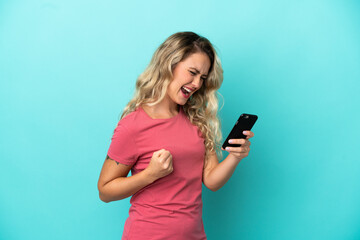 Fototapeta premium Young Brazilian woman isolated on blue background using mobile phone and doing victory gesture