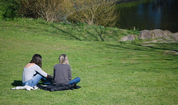 Duas raparigas a conversar sentadas na erva de um parque viradas para um lago em dia de sol