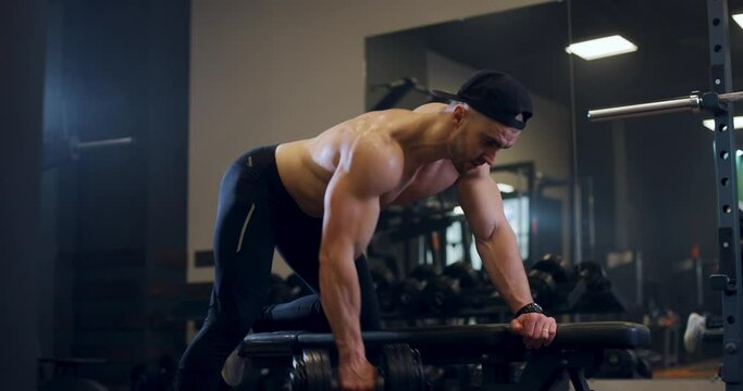Handsome shirtless sporty muscular bodybuilder man excercise with weights building his back muscle in professional sport gym. Bodybuilder lifts dumbbell on bench, slow motion