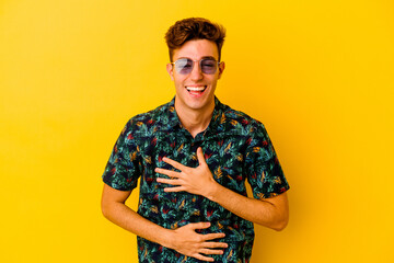 Young caucasian man wearing a Hawaiian shirt isolated on yellow background laughs happily and has fun keeping hands on stomach.