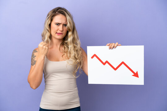 Young Brazilian woman isolated on purple background holding a sign with a decreasing statistics arrow symbol and angry