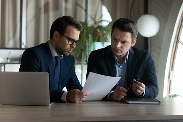 Concentrated male employees colleagues sit at desk in office brainstorm over financial paperwork. Serious businessmen consider paper document at meeting, cooperate at workplace. Teamwork concept.